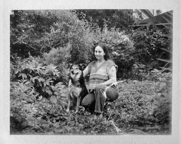 Black and white photo of a woman with long brown hair in a knitted vest, kneeling next to her dog.